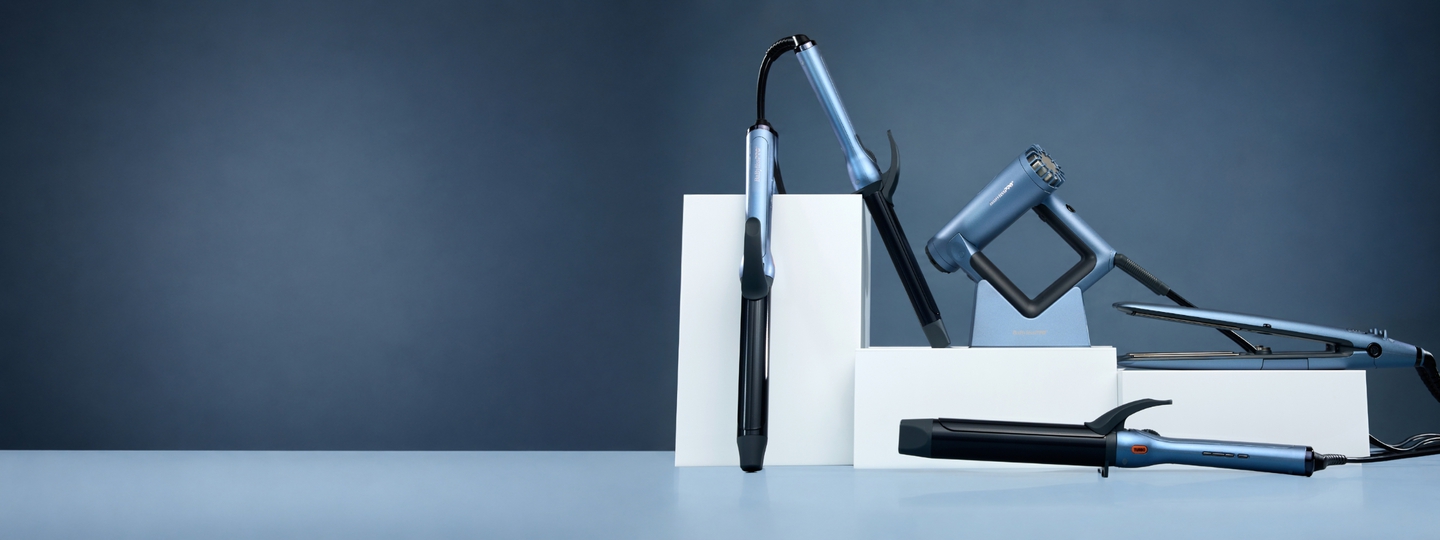 collection of styling tools featuring a flat iron, three curling irons, and a square-shaped hair dryer, on white blocks in front of a grey and blue background